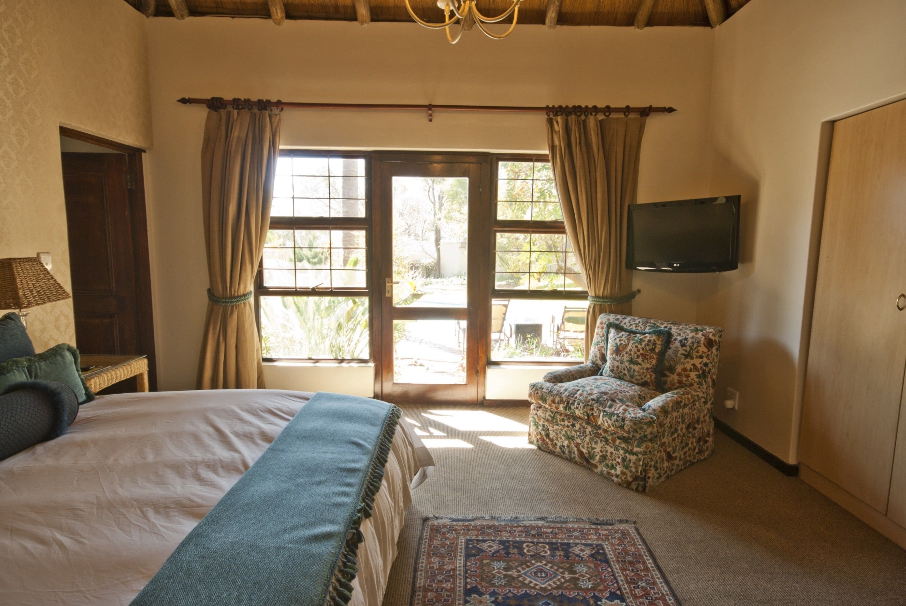 A bedroom with a large bed, a patterned armchair, a television, and a view through a glass door.