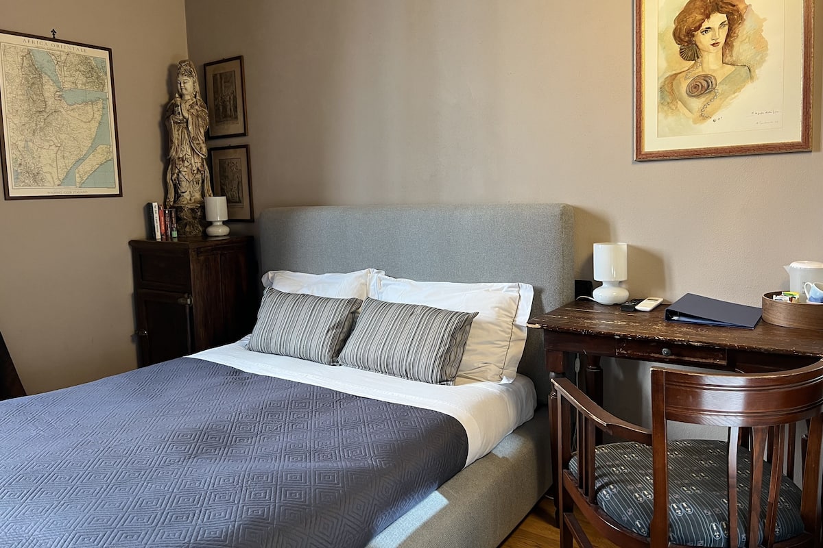 A neatly made bed with a patterned bedspread, a grey upholstered headboard, and a wooden nightstand with a lamp and a book.