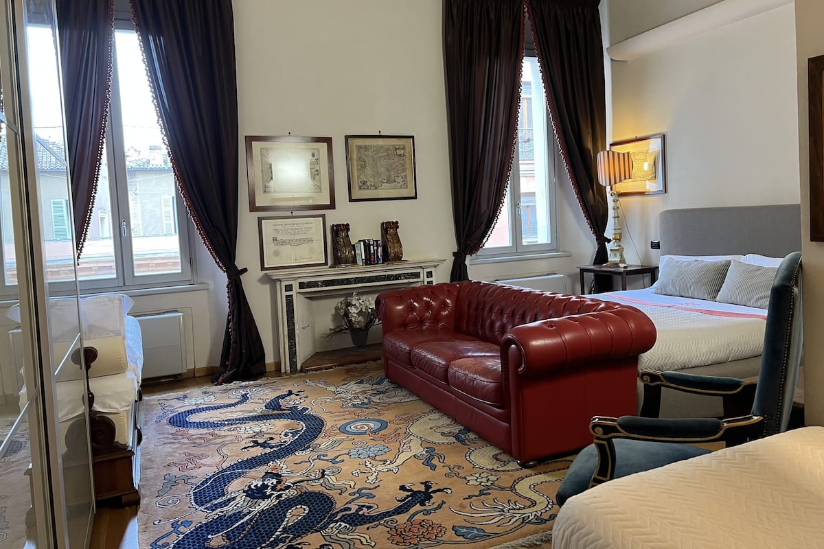 A hotel room with a red leather sofa, a bed with a grey headboard, a blue armchair, a patterned rug, and two framed certificates on the wall.