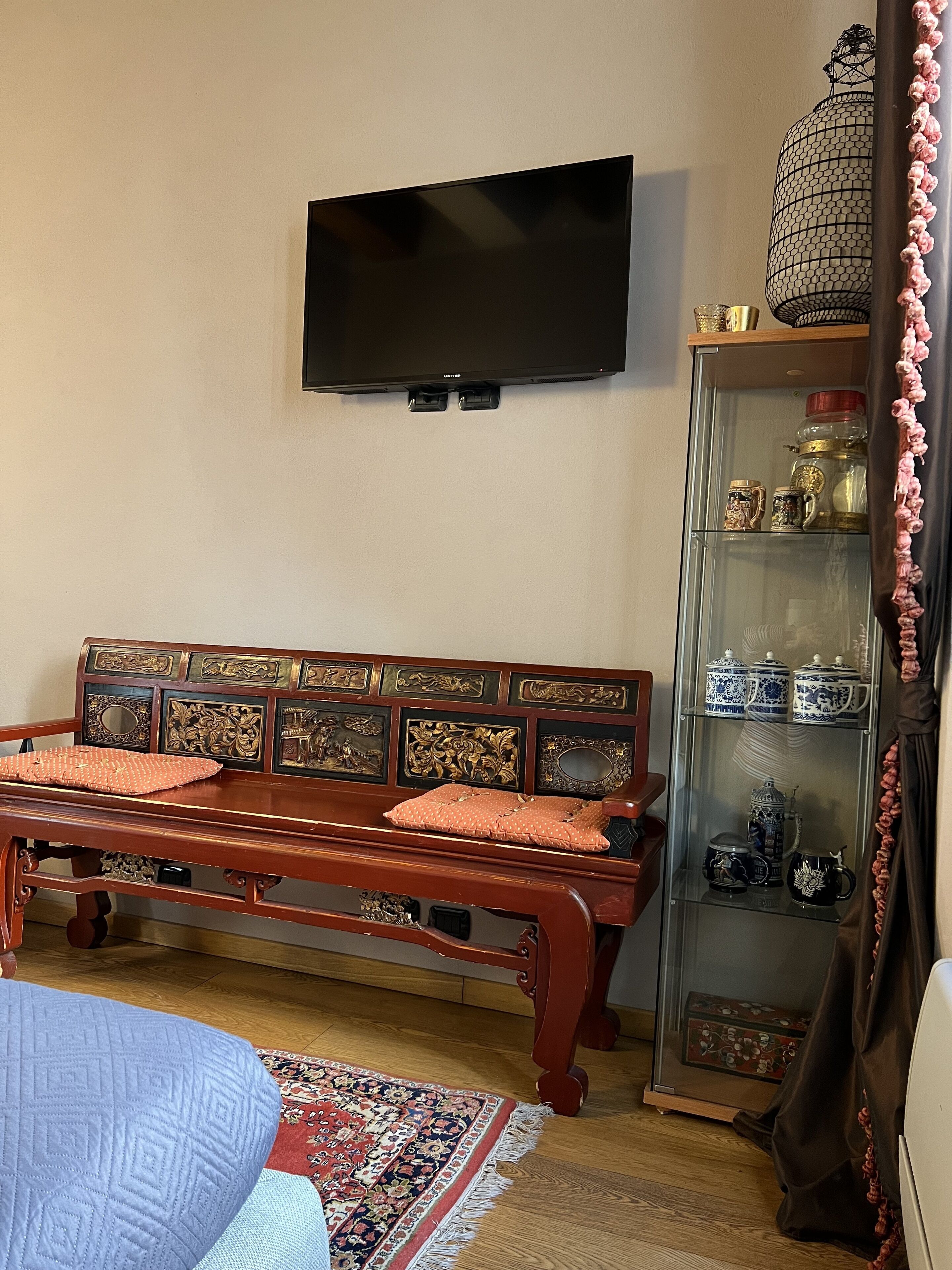 Una sala de estar con un sofá de madera, un televisor montado en la pared, un aparador de vidrio con objetos decorativos y una alfombra estampada en el suelo.