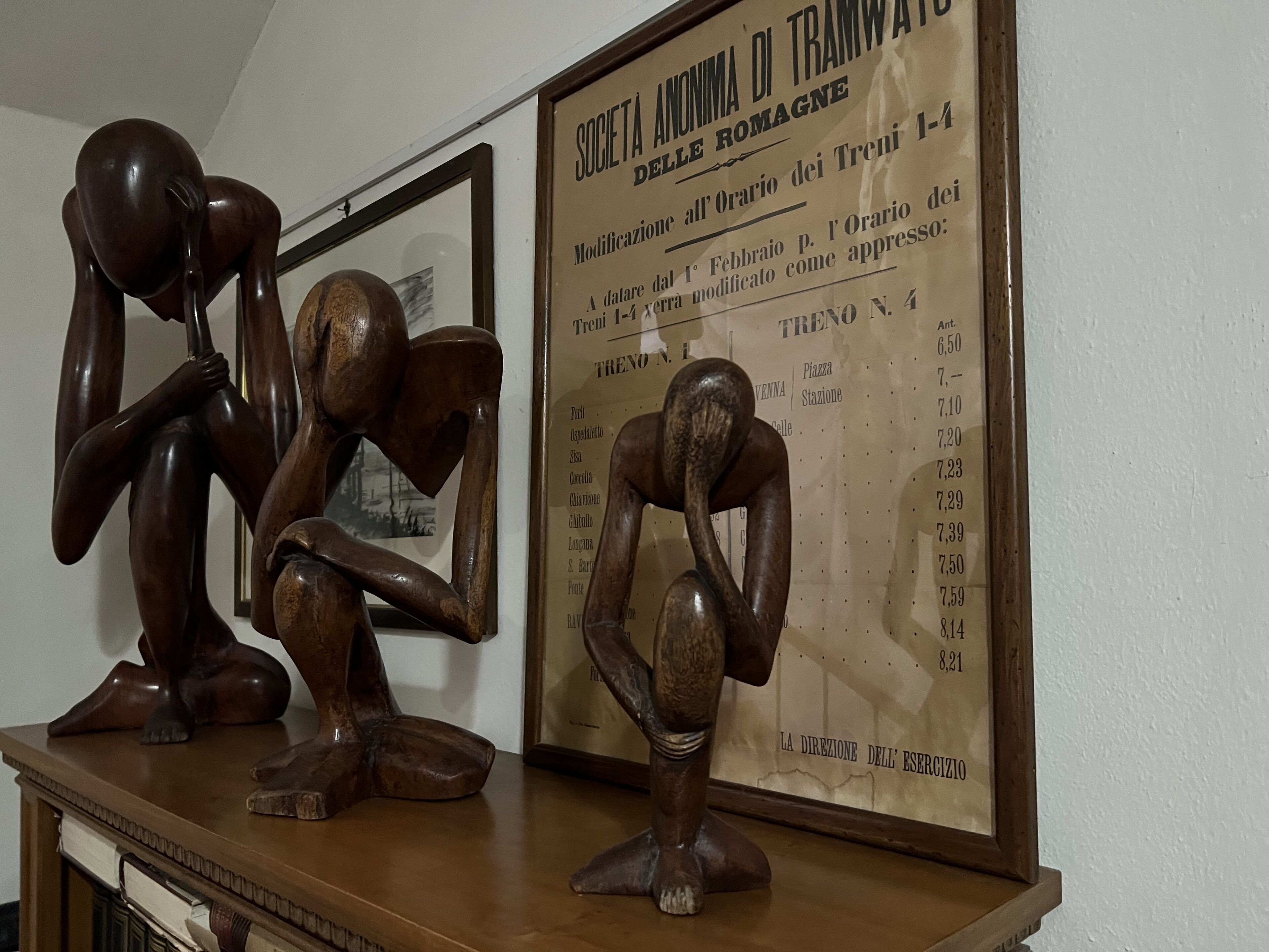 A wooden sculpture of a figure in a contemplative pose on a table, with a framed sign listing room numbers and directions in the background.