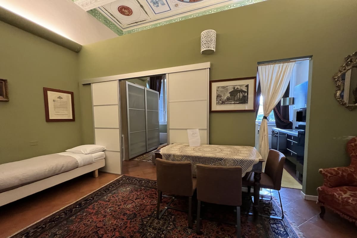 A room with a bed, a dining table with chairs, a patterned rug, a wardrobe, and a kitchen visible through an open door.