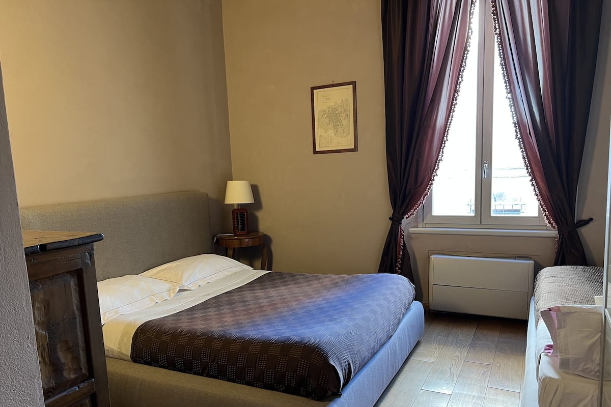 A hotel room with a bed, a bedside table with a lamp, a window with curtains, and a radiator.