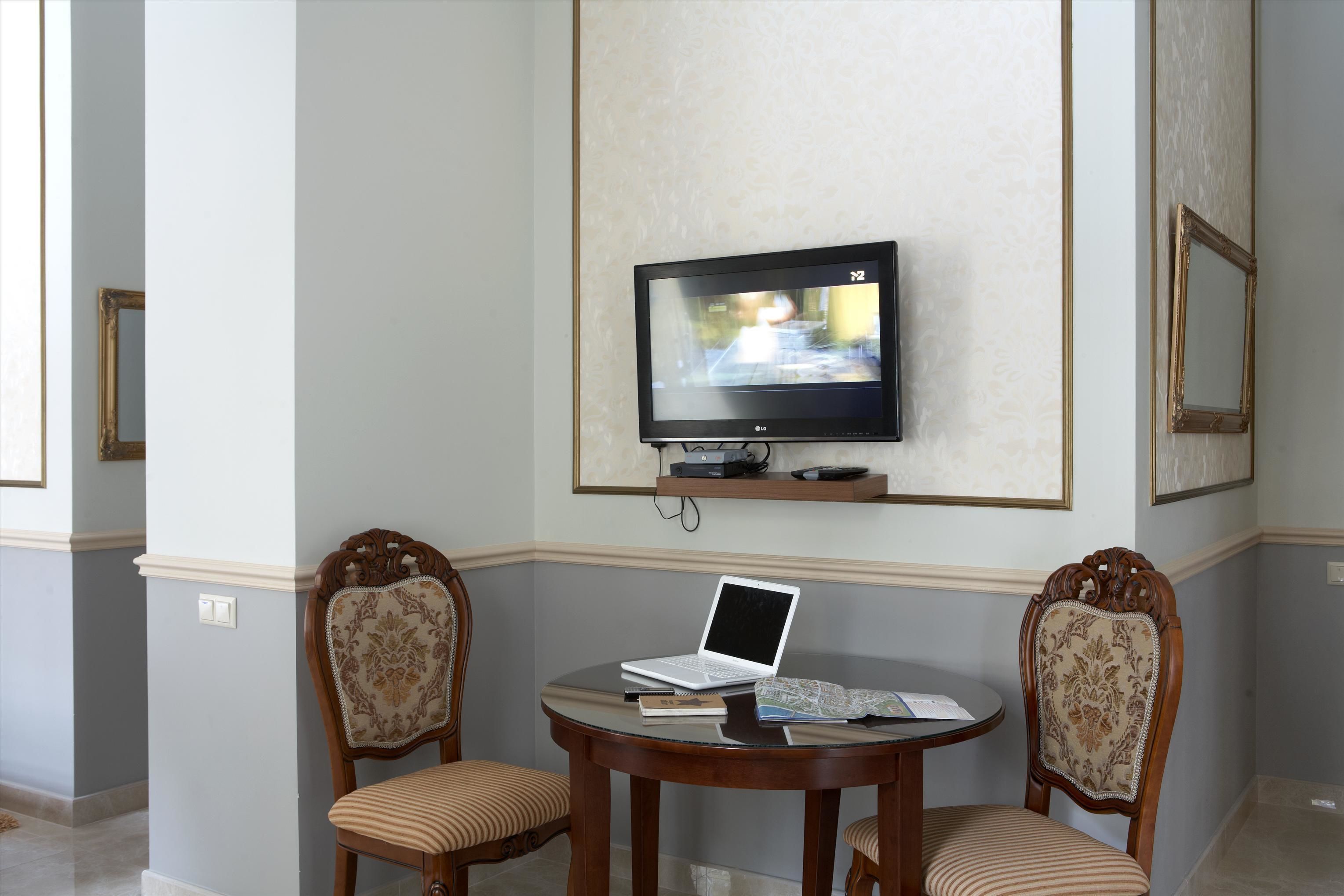 A room with a TV mounted on the wall, a laptop on a small table, two upholstered chairs, and a framed picture on the wall.