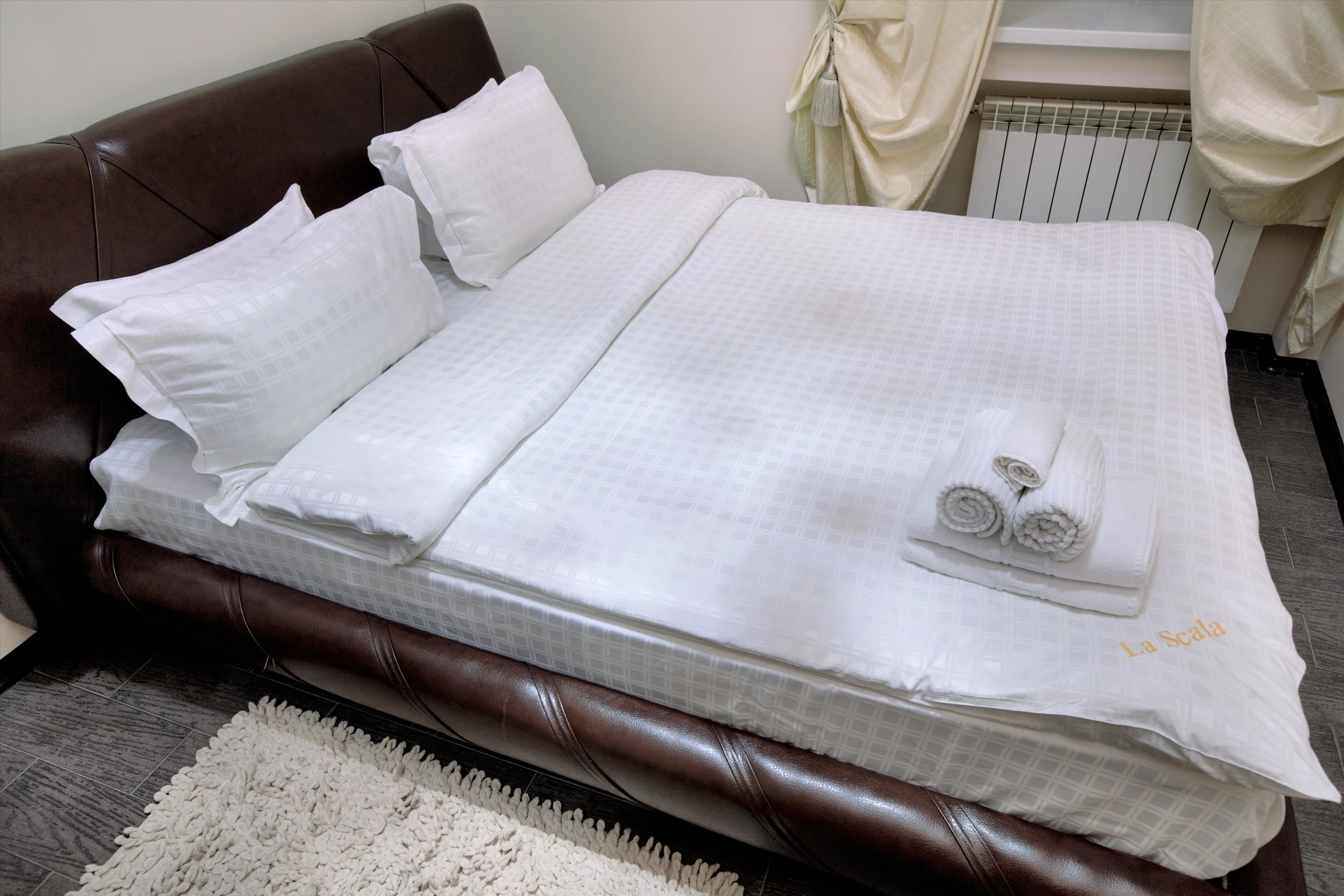 A neatly made bed with white linens and a rolled towel on top, a brown leather headboard, and a white rug on the floor.