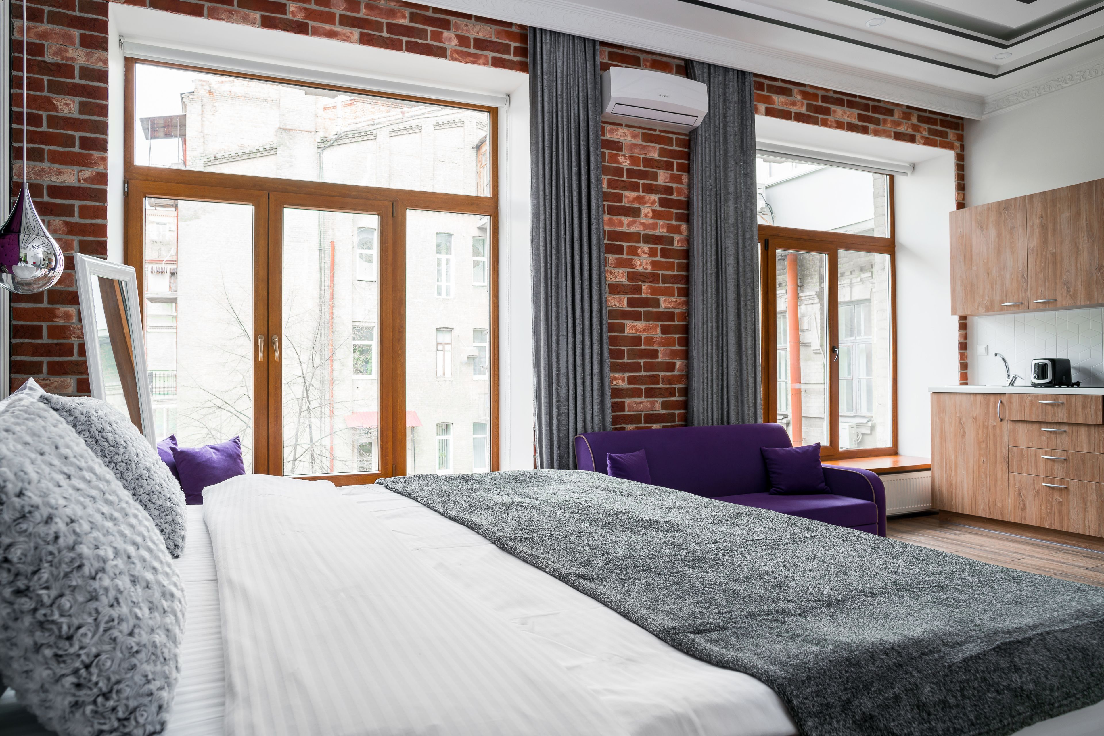 Habitación de hotel moderna con una cama grande, un sofá púrpura, una zona de cocina y ventanas amplias con una pared de ladrillo.