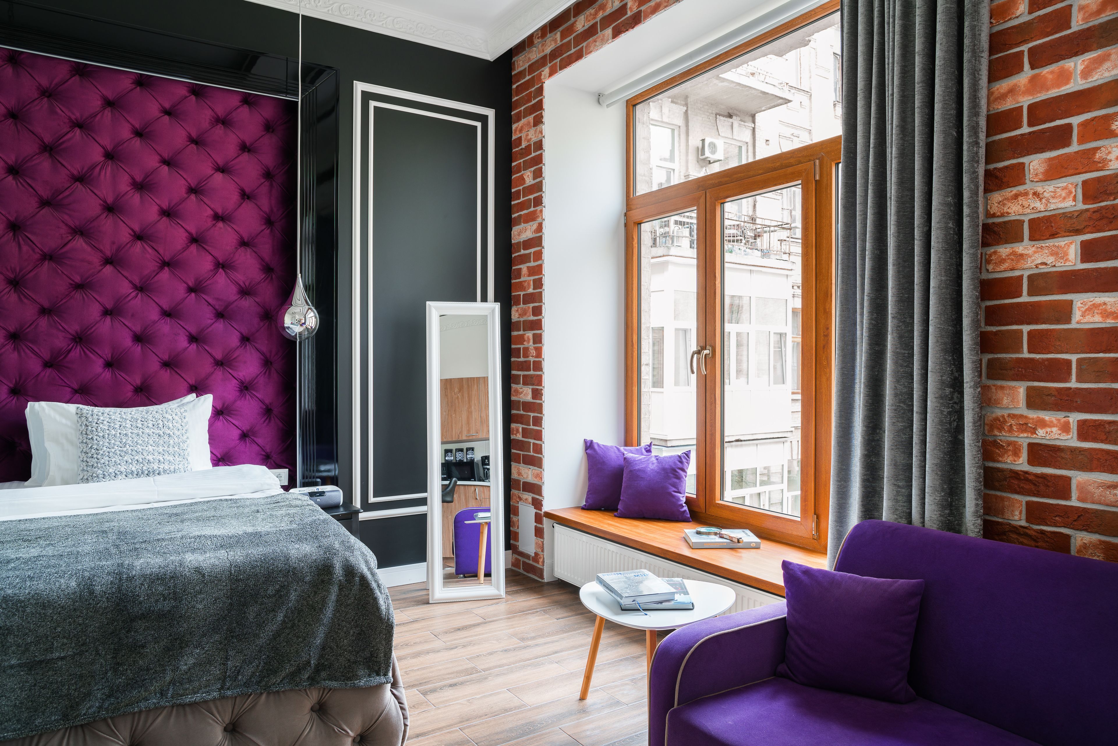A modern hotel room with a tufted headboard, a large window with curtains, a wooden bench, and a purple sofa.