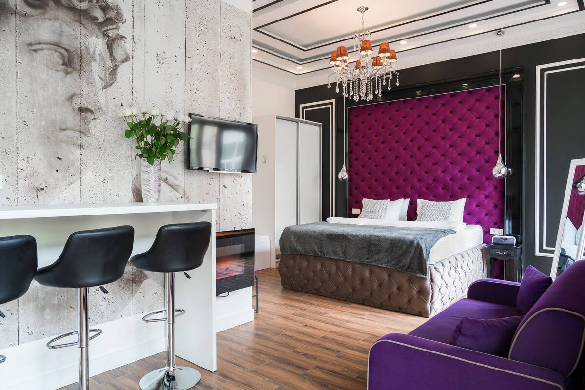 Una habitación de hotel moderna con cama, televisión, zona de bar con taburetes y un sofá púrpura.