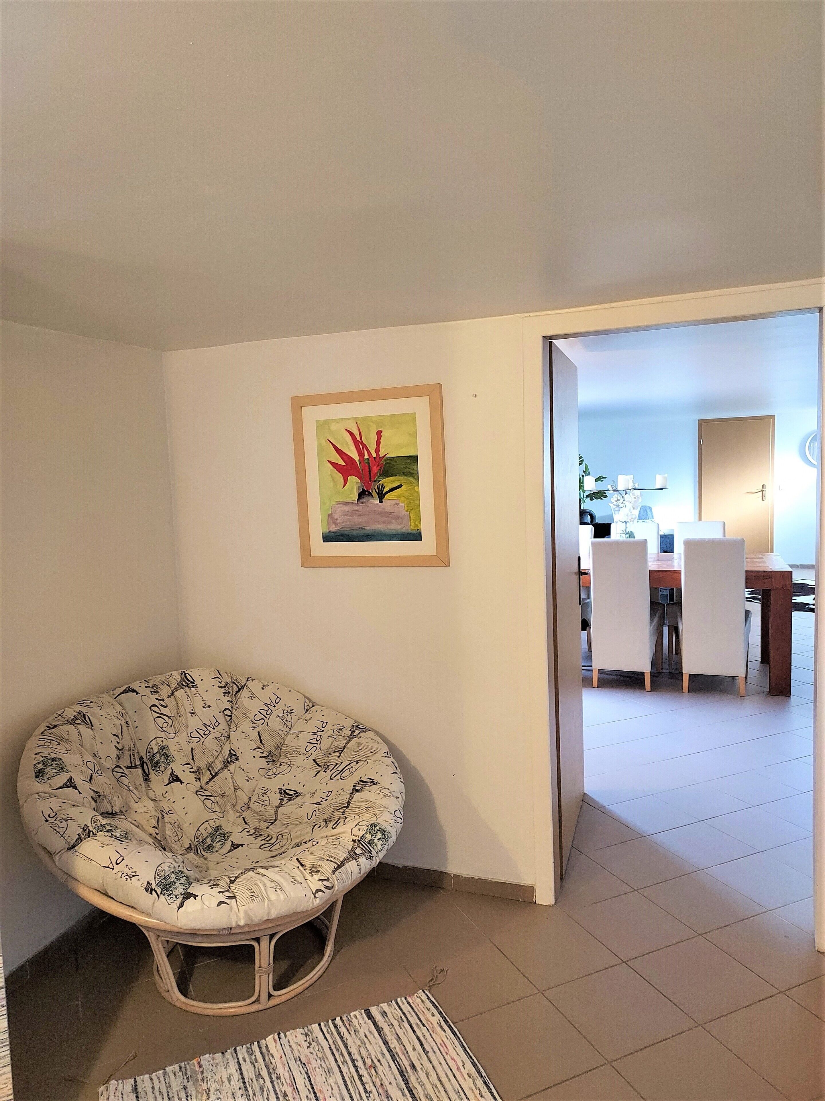 Habitación de esquina con una silla redonda y estampada, un cuadro enmarcado, y vista a otro cuarto con zona de comedor.