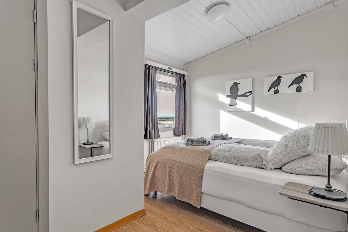 A modern bedroom with a bed, a bedside table with a lamp, a window with curtains, and artwork on the wall.