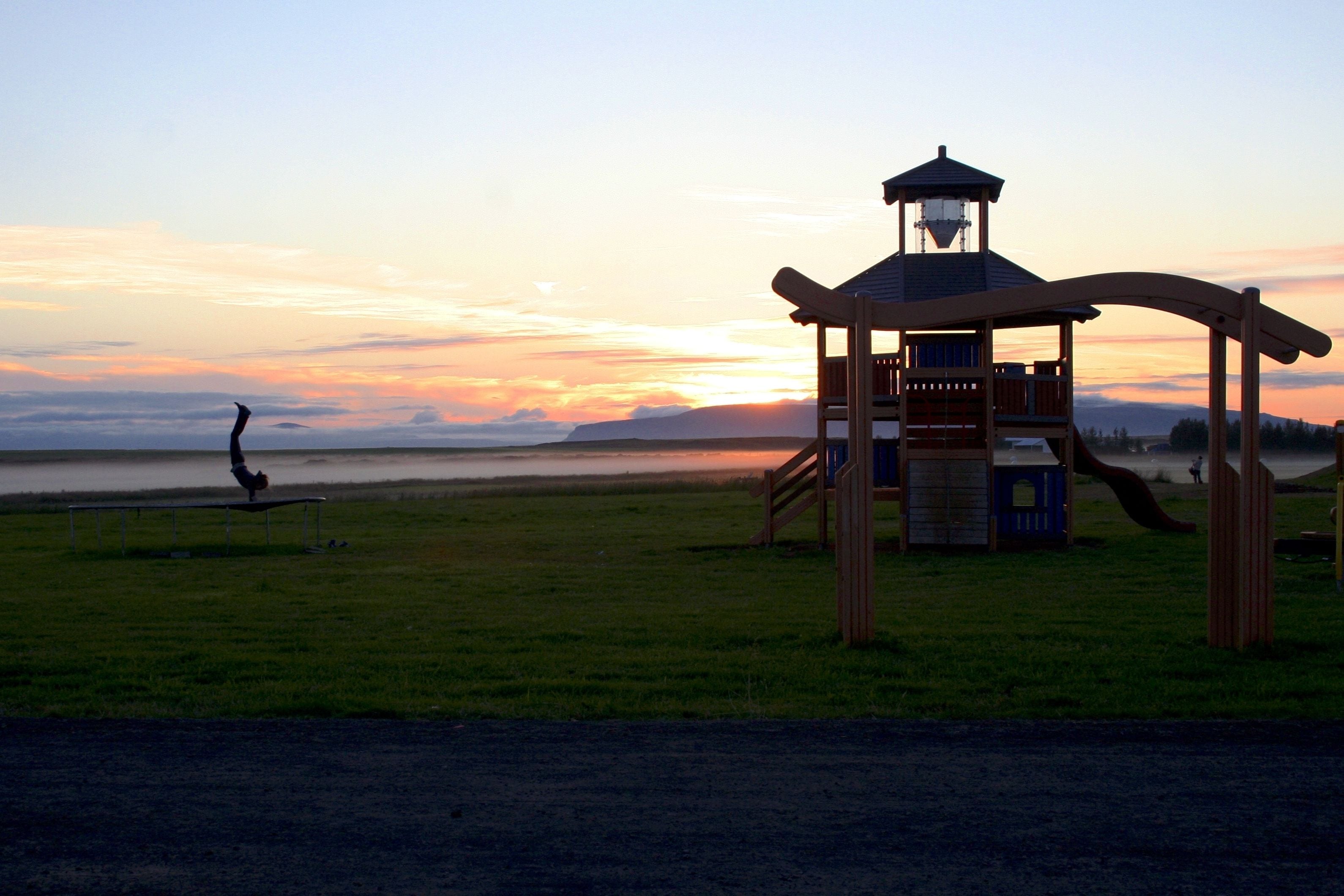 A playground with a wooden structure and slides, set against a sunset over a body of water.