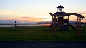 A playground with a wooden structure and slides, set against a sunset over a body of water.