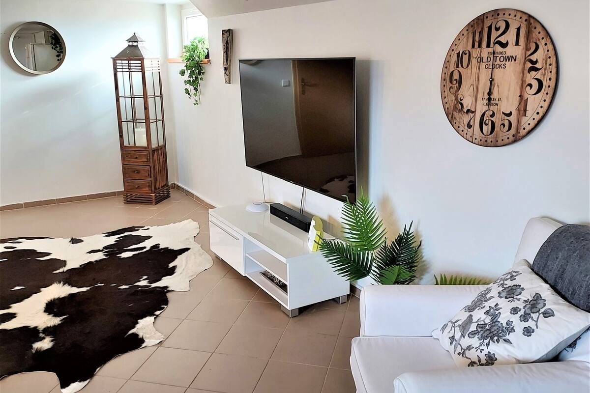 A modern living room with a flat-screen TV, a wooden clock, a cowhide rug, and a white sofa with floral pillows.
