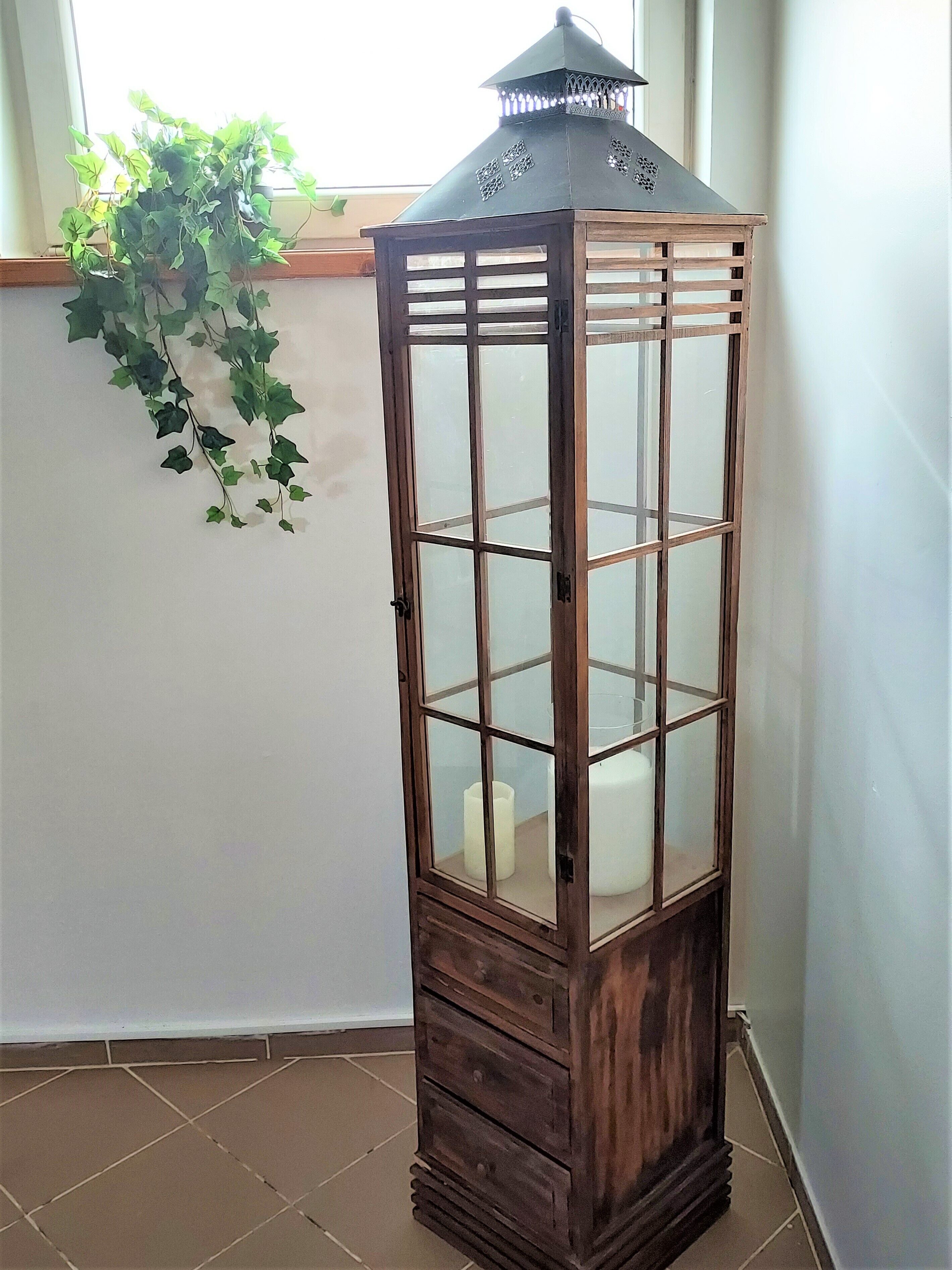 A tall, narrow wooden cabinet with glass doors and drawers, placed in a corner with a window and a hanging plant in the background.