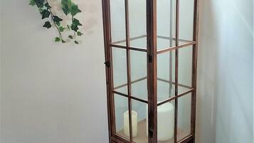 A tall, narrow wooden cabinet with glass doors and drawers, placed in a corner with a window and a hanging plant in the background.