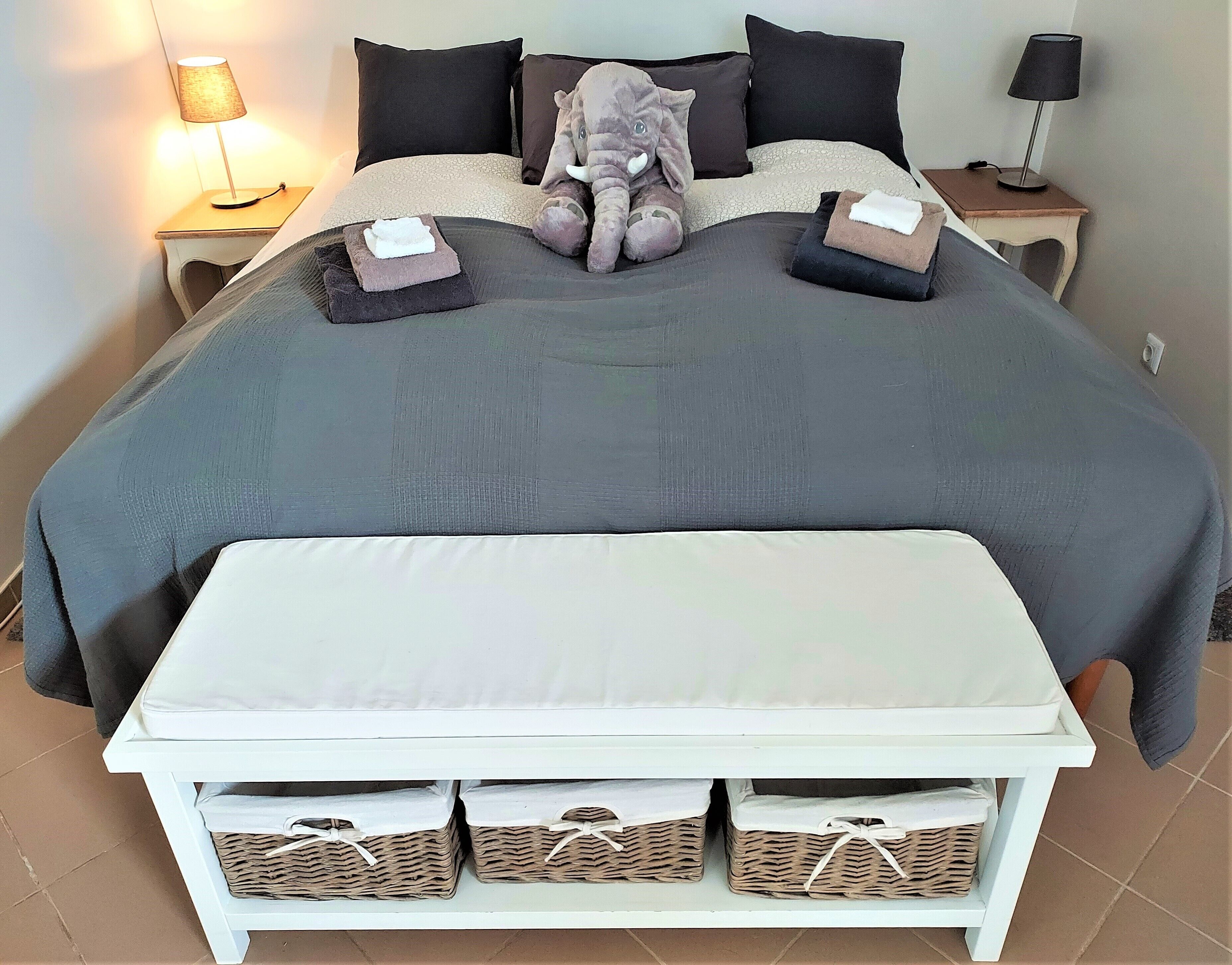 Una cama bien tendida con un edredón gris, dos almohadas, un peluche de elefante y una lámpara de noche a cada lado.