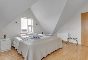 Un dormitorio con techo inclinado, una cama con cubrecama blanco, una mesita de noche, un radiador y una ventana con vistas.