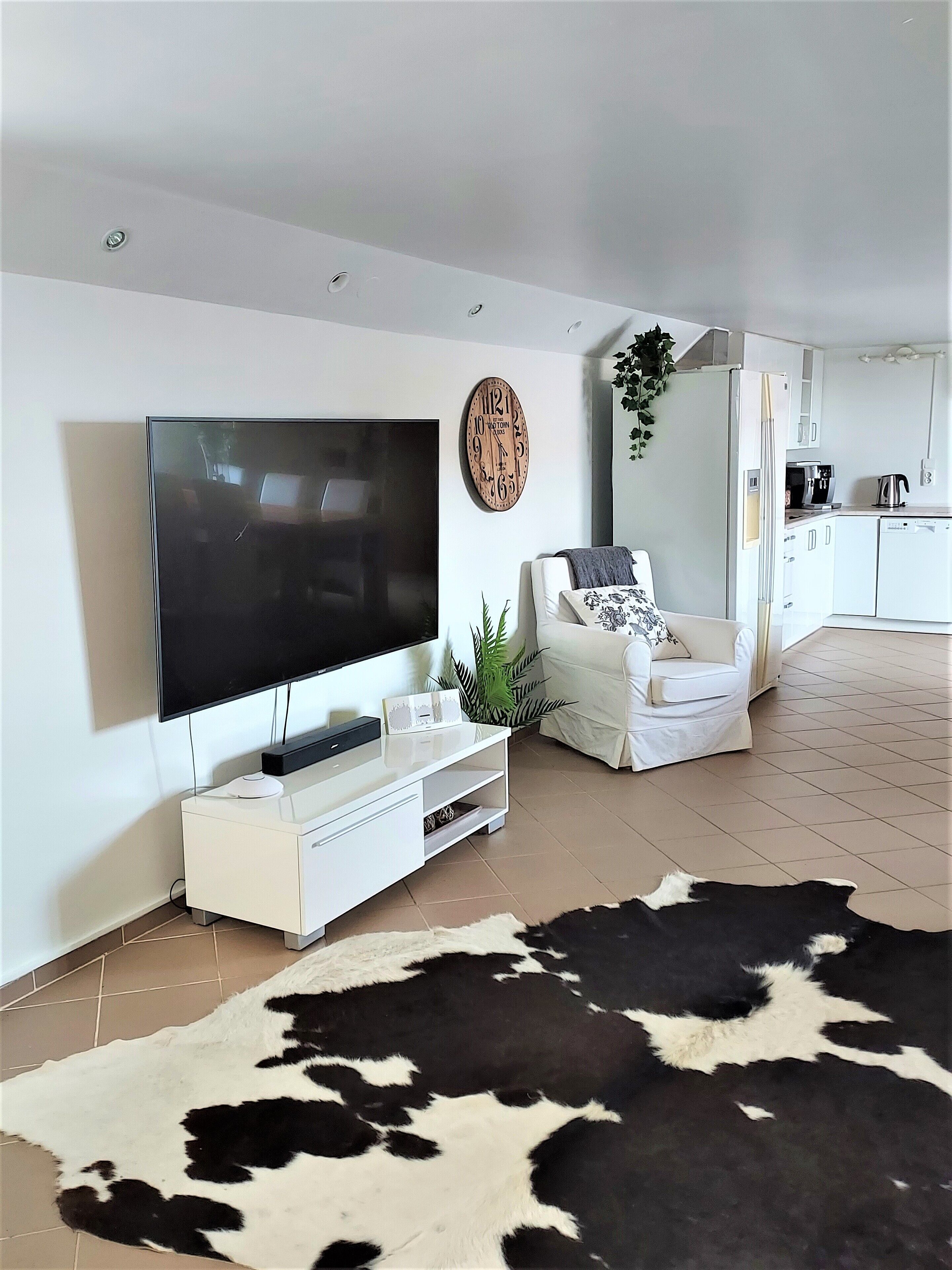 A modern living room with a flat-screen TV, a white sofa, a cowhide rug, and a kitchen in the background.