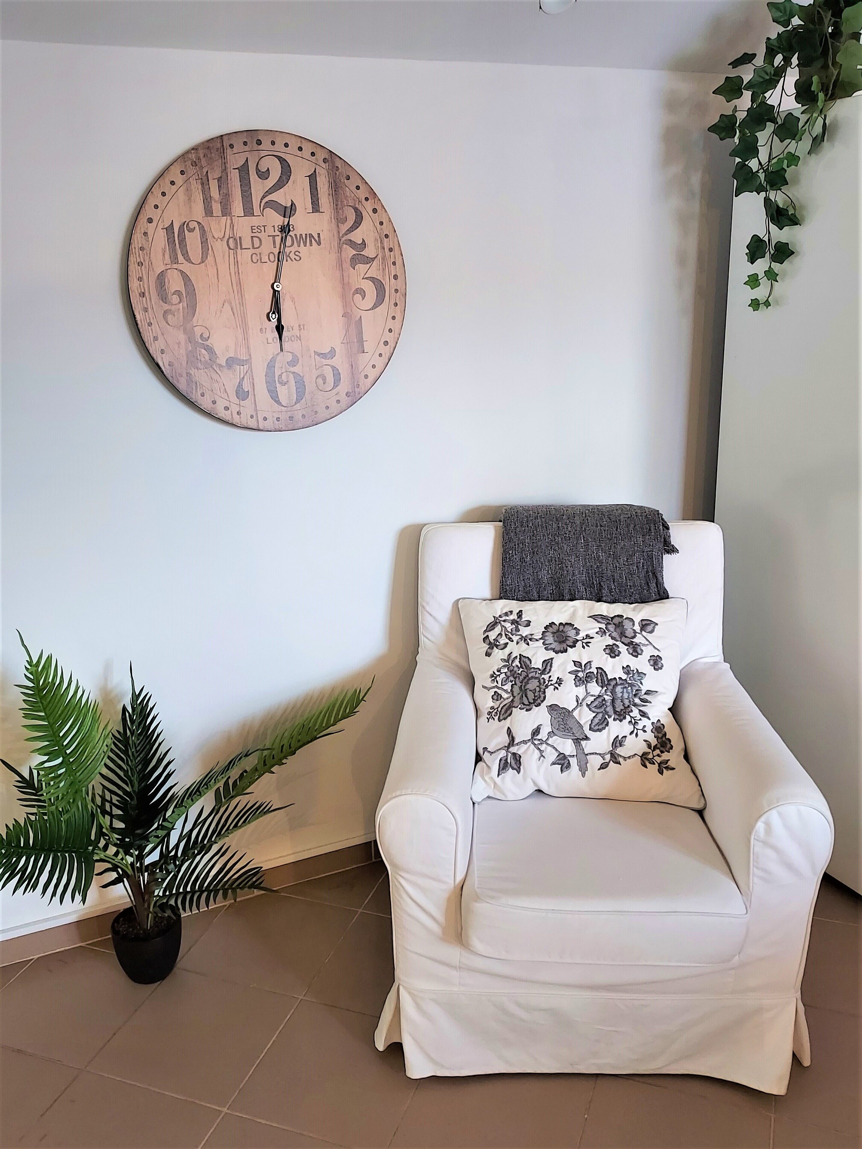 A white armchair with a floral pillow and a grey throw blanket, a round wooden clock with numbers, a potted plant, and a wall with a plant hanging on it.