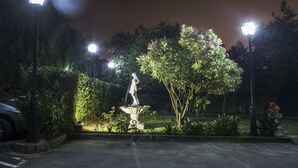 A nighttime garden with a statue, illuminated by street lamps, surrounded by greenery and a paved area.