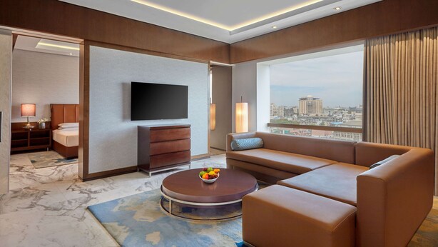 A modern hotel room with a large window, a flat-screen TV, a sofa, a coffee table with fruit, and a bed with a nightstand.