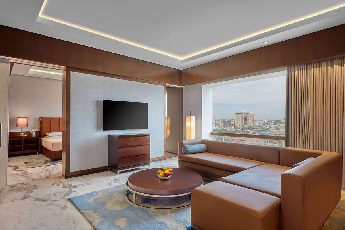 A modern hotel room with a large window, a flat-screen TV, a sofa, a coffee table with fruit, and a bed with a nightstand.