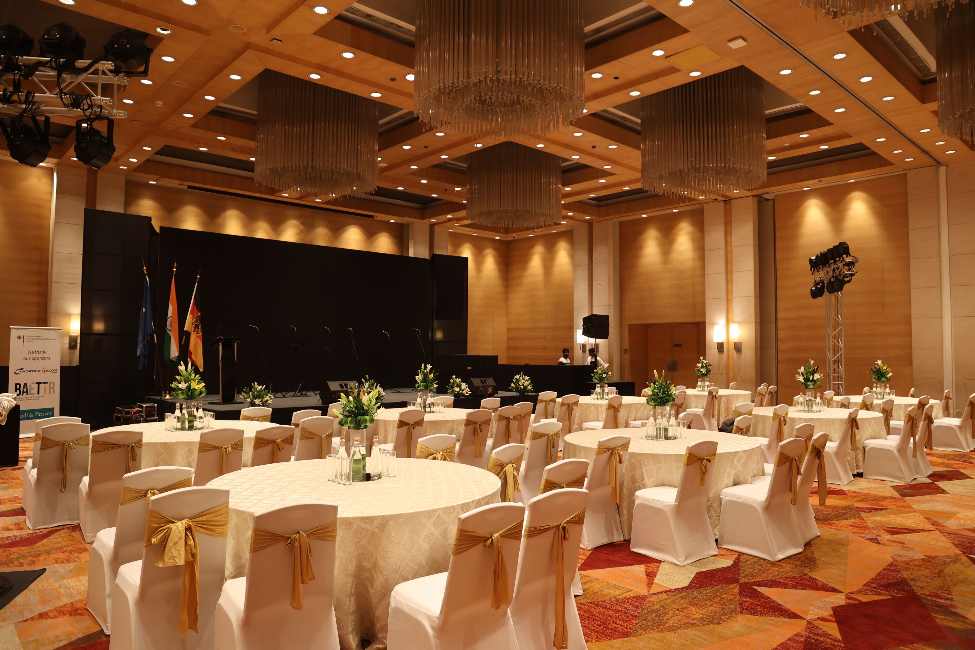 Ein Festsaal mit runden Tischen, weißen Stühlen mit goldenen Bändern und einer Bühne mit Flaggen.
