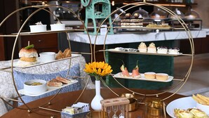 A three-tiered dessert display with various pastries and cakes, a vase with yellow flowers, and a teapot on a wooden table.