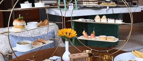 A three-tiered dessert display with various pastries and cakes, a vase with yellow flowers, and a teapot on a wooden table.