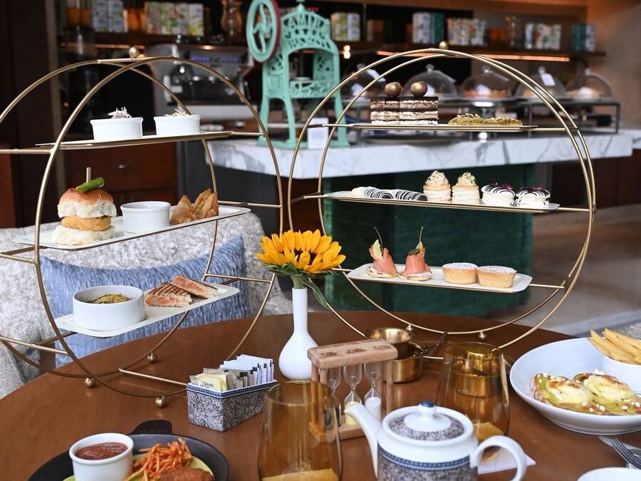 A three-tiered dessert display with various pastries and cakes, a vase with yellow flowers, and a teapot on a wooden table.