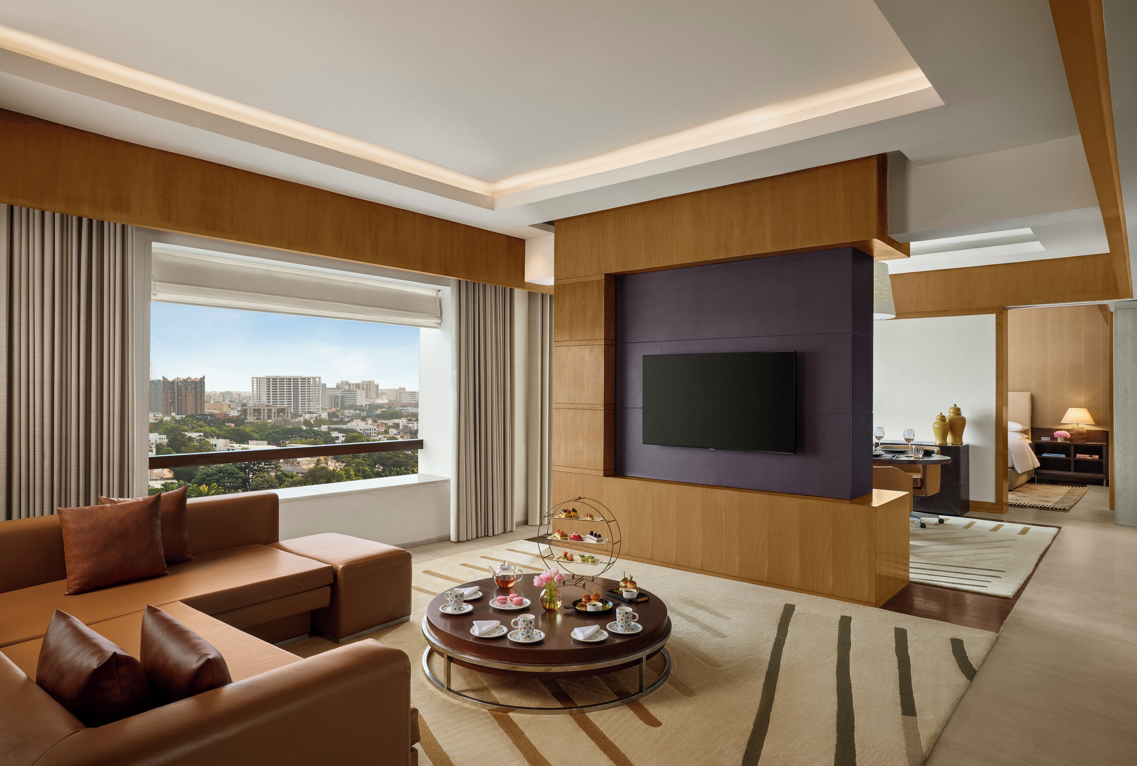 Habitación de hotel moderna con un ventanal grande, un televisor de pantalla plana, una mesa de centro redonda con aperitivos y una zona de estar con sofás de cuero.