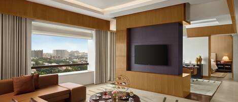 Habitación de hotel moderna con un ventanal grande, un televisor de pantalla plana, una mesa de centro redonda con aperitivos y una zona de estar con sofás de cuero.