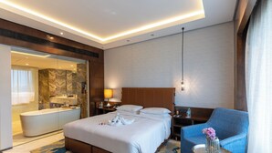 A hotel room with a large bed, a sitting area with a chair, a small table, a bathroom with a bathtub, and a large mirror.