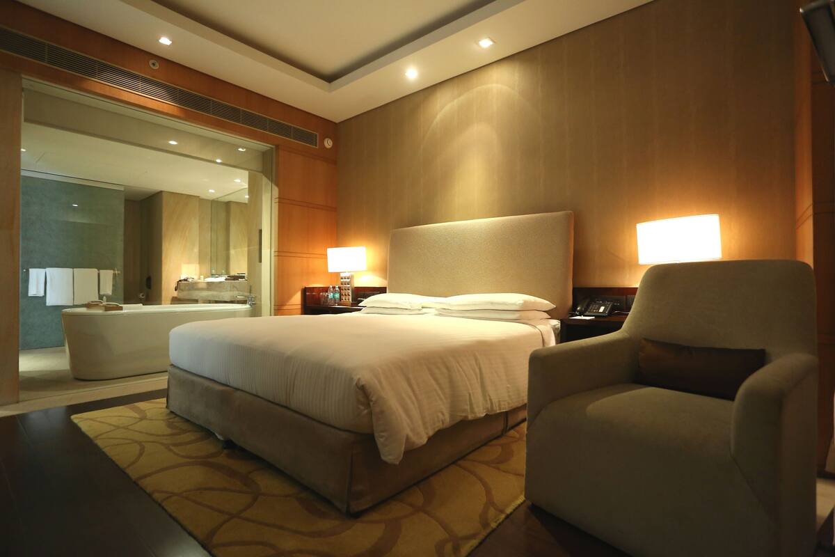 A hotel room with a large bed, a sitting area with a chair, a bedside table with a lamp, and a large mirror.