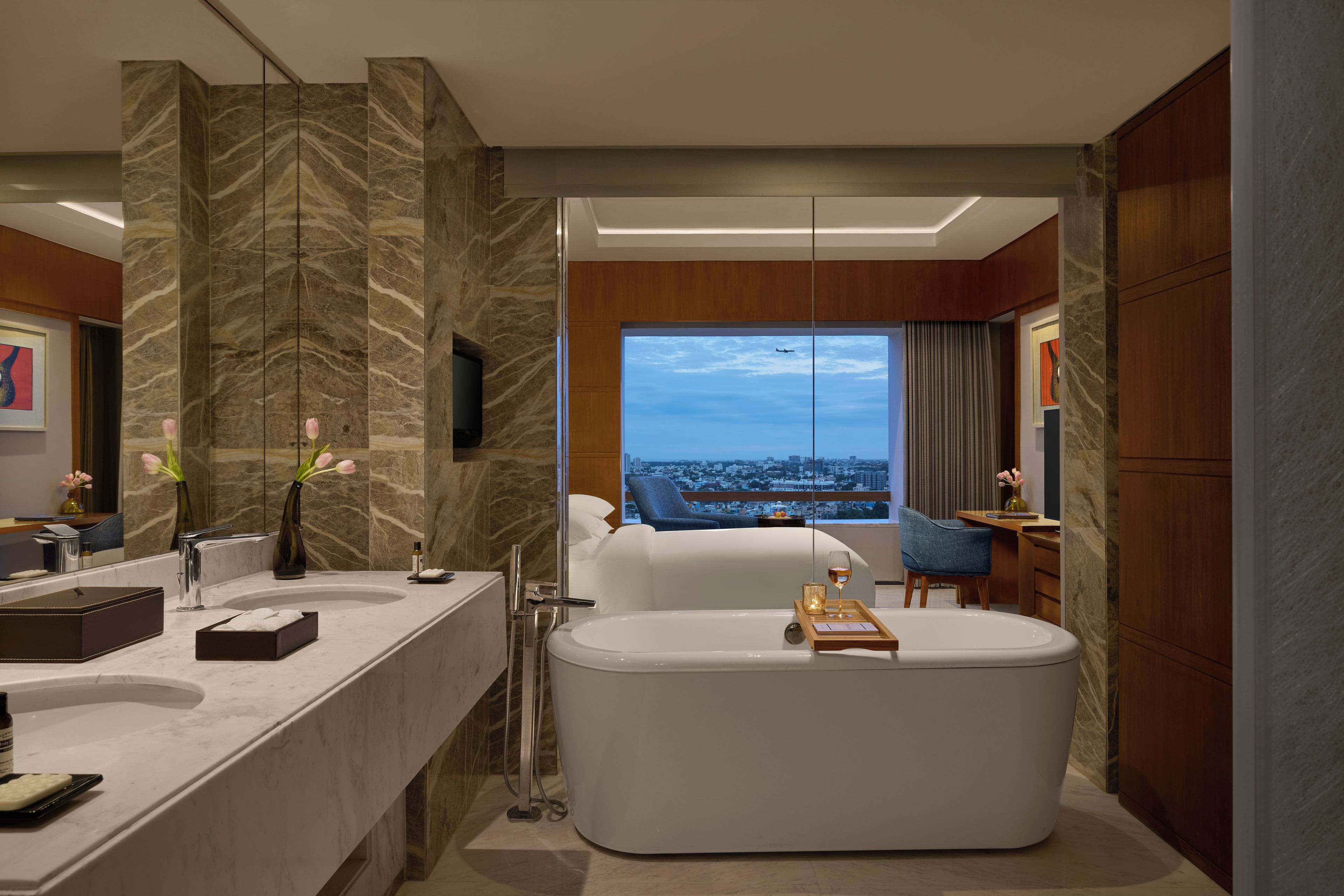 A modern bathroom with a large bathtub, a vanity with a mirror, and a view of a cityscape through a large window.