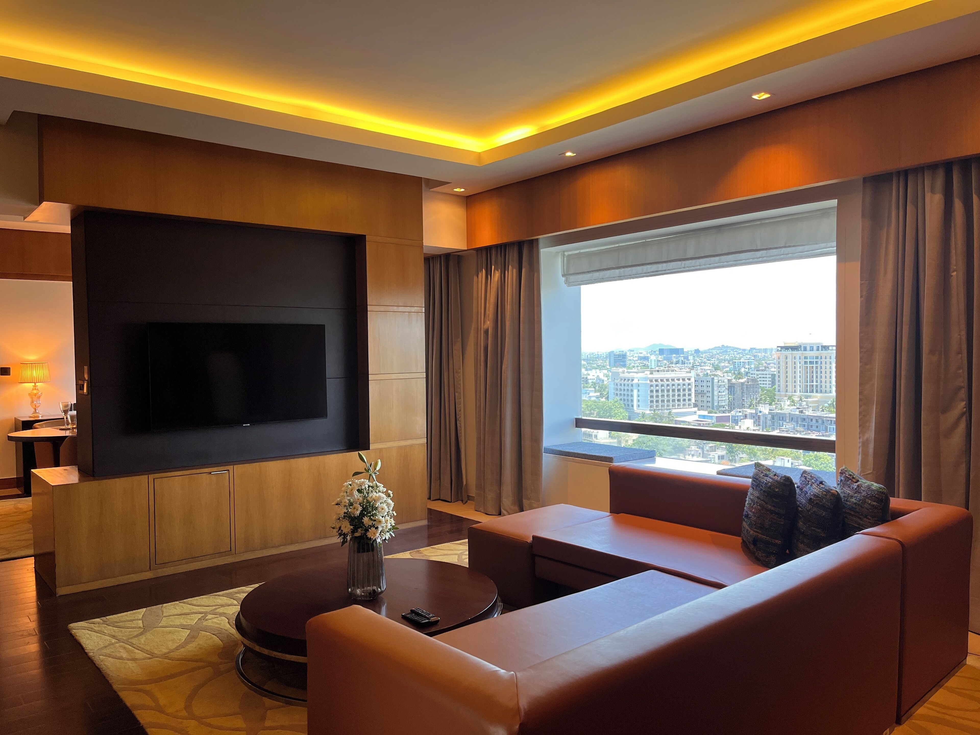Een moderne woonkamer met een flatscreen televisie aan de muur, een bank, een salontafel en een groot raam met uitzicht op de stad.