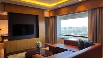 A modern living room with a flat-screen TV mounted on the wall, a sofa, a coffee table, and a large window offering a city view.