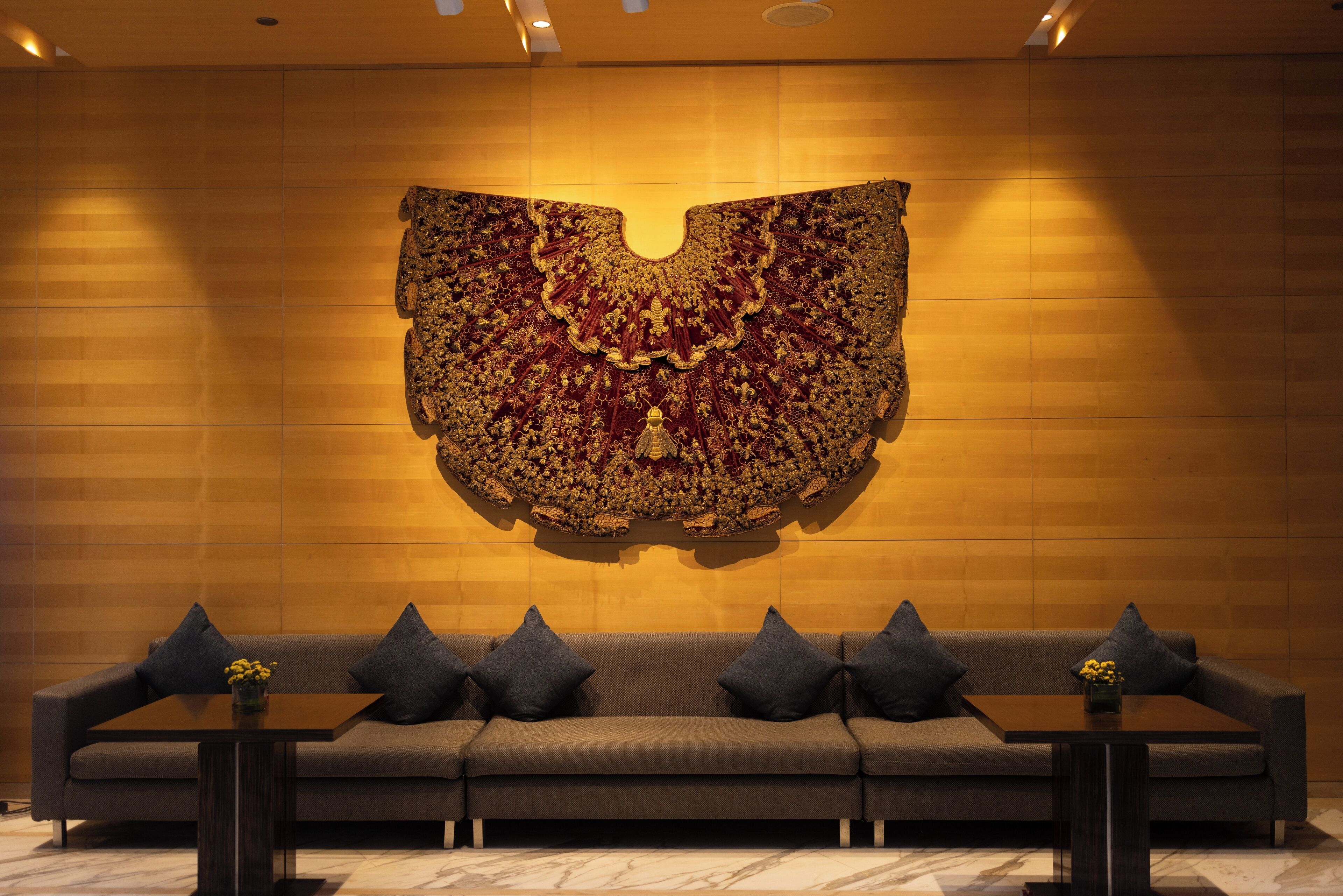 Een moderne lobby met een groot, versierd wandtapijt, een grijze bank met kussens en twee kleine tafels met vazen.