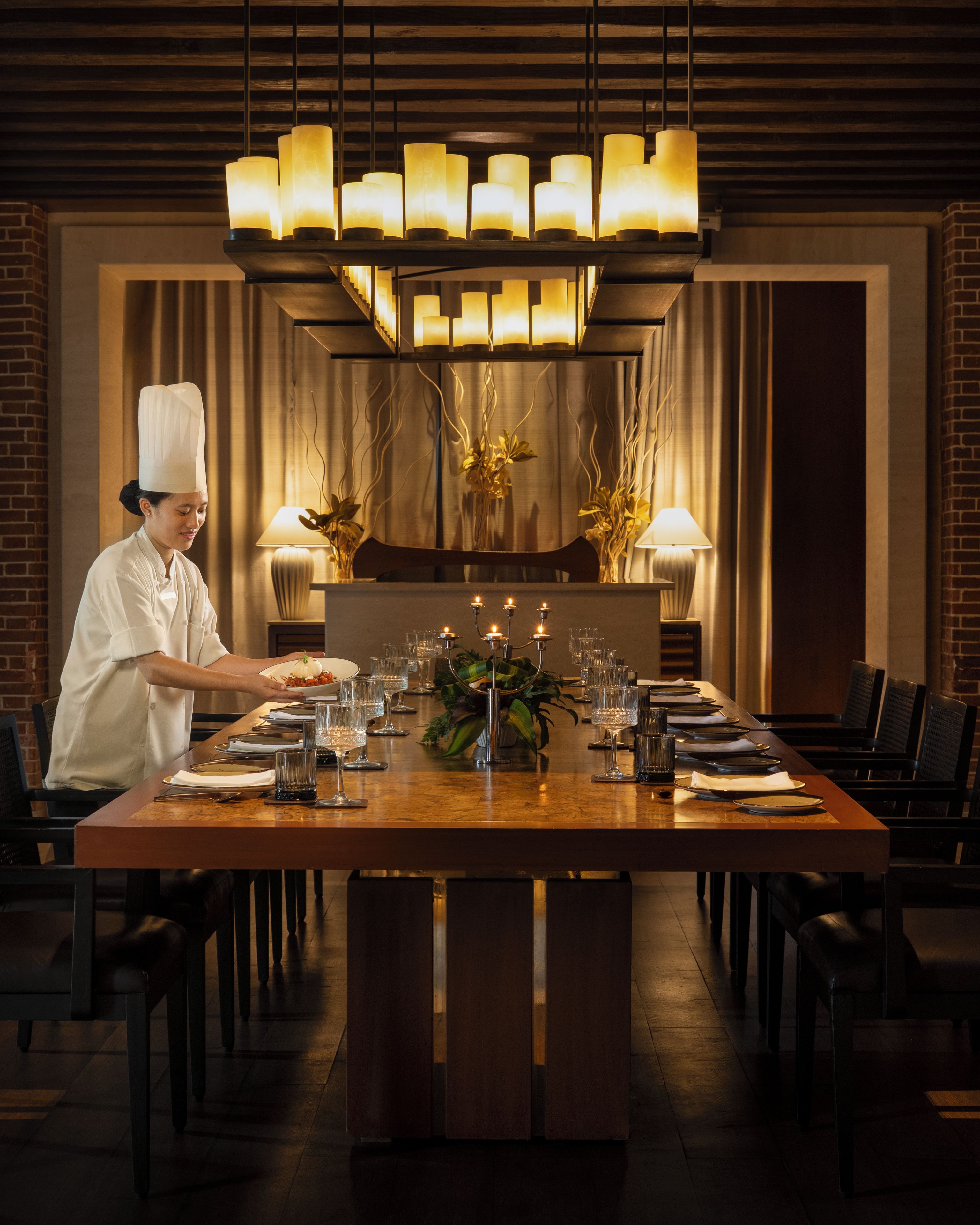 A restaurant setting with a chef preparing a dish, a long dining table set with chairs, and a modern chandelier.