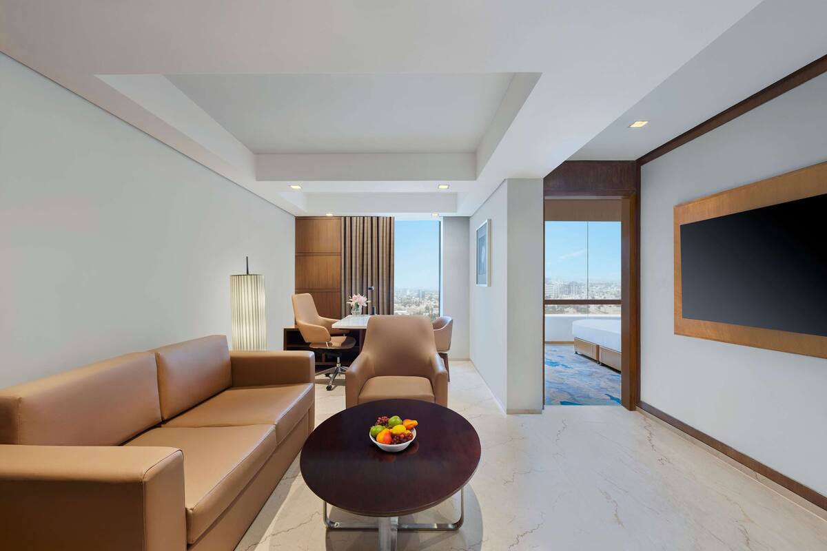 A modern living room with a sofa, a coffee table with fruit, a dining area with chairs, and a large TV on the wall.
