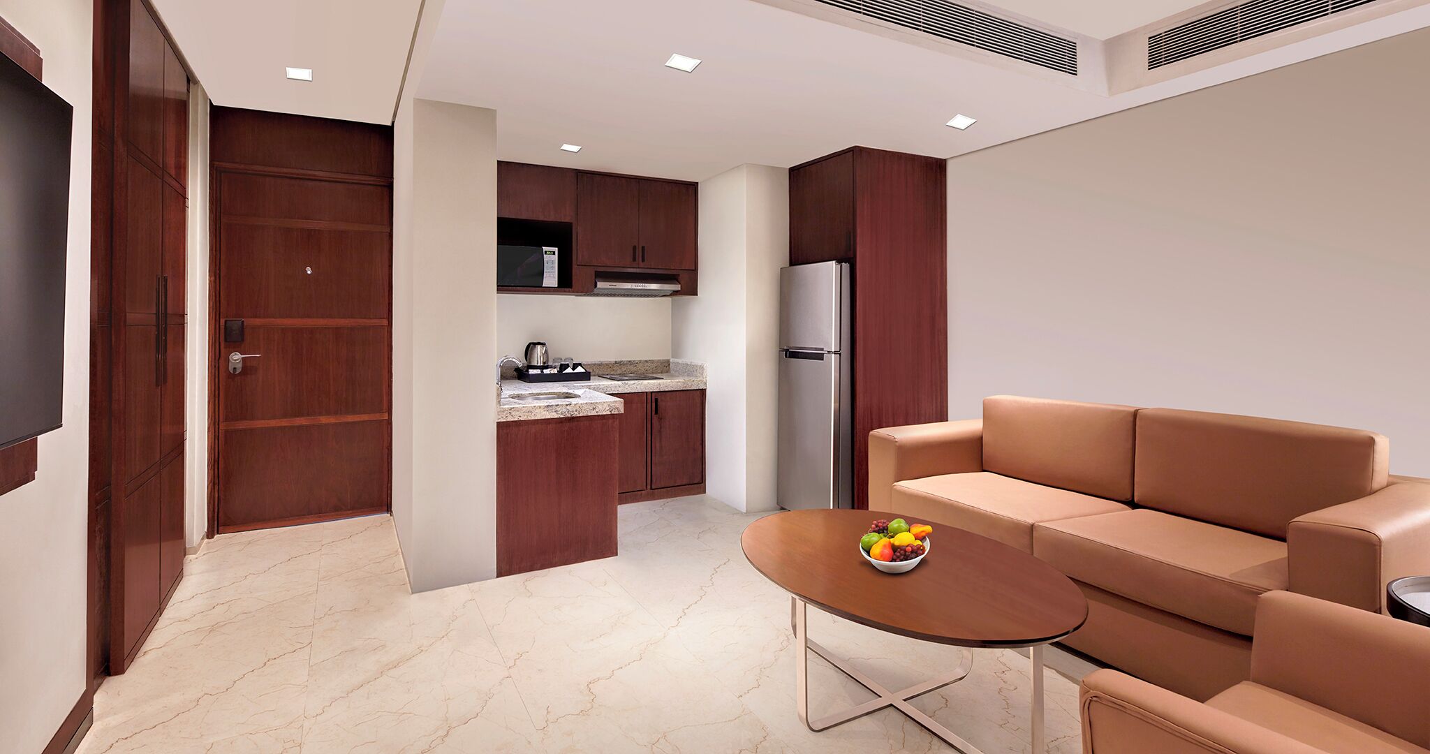 Cocina moderna con armarios de madera oscura, un refrigerador de acero inoxidable y una encimera de mármol. En la sala de estar, hay un sofá color beige y una mesita con un tazón de fruta.
