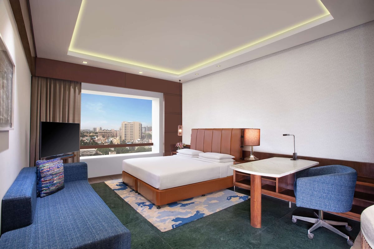 A hotel room with a bed, a desk, a chair, a television, and a large window offering a city view.