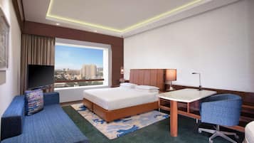 Habitación de hotel con cama, escritorio, silla, televisor y ventana grande con vistas a la ciudad.