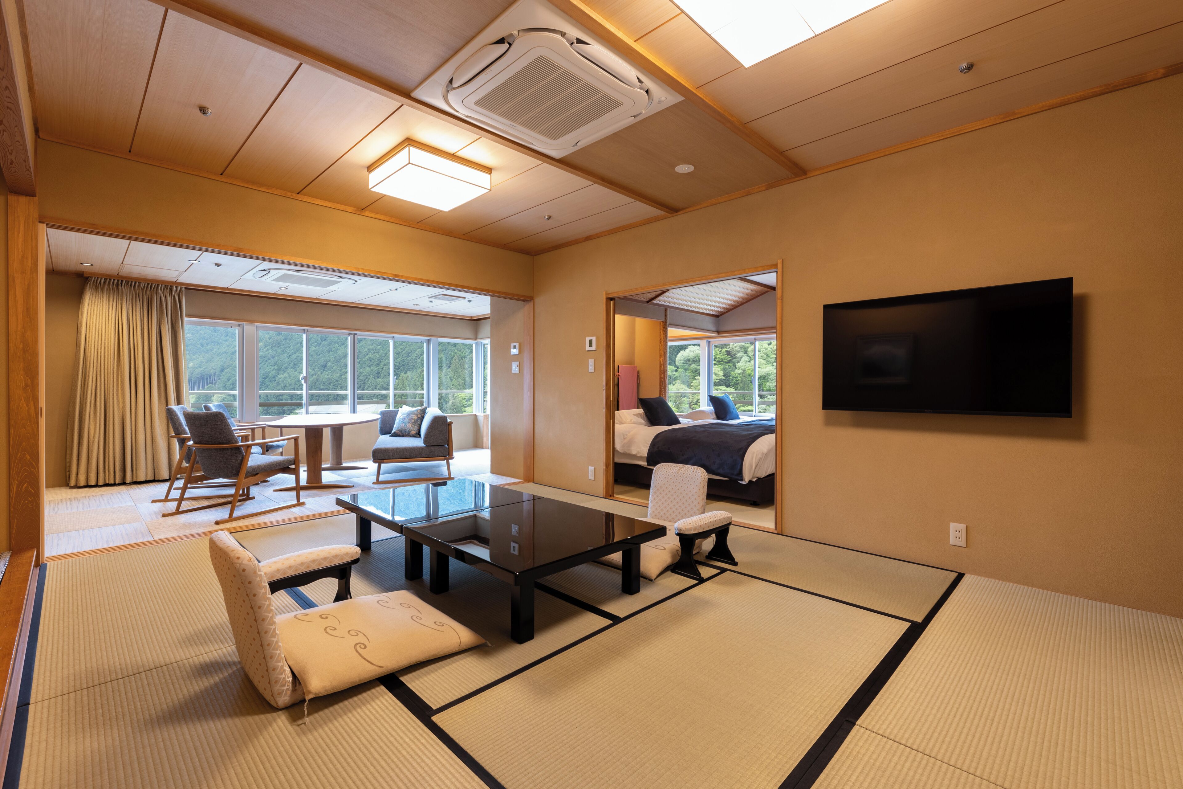 A traditional Japanese-style room with tatami flooring, a low table, chairs, a bed, and a TV mounted on the wall.