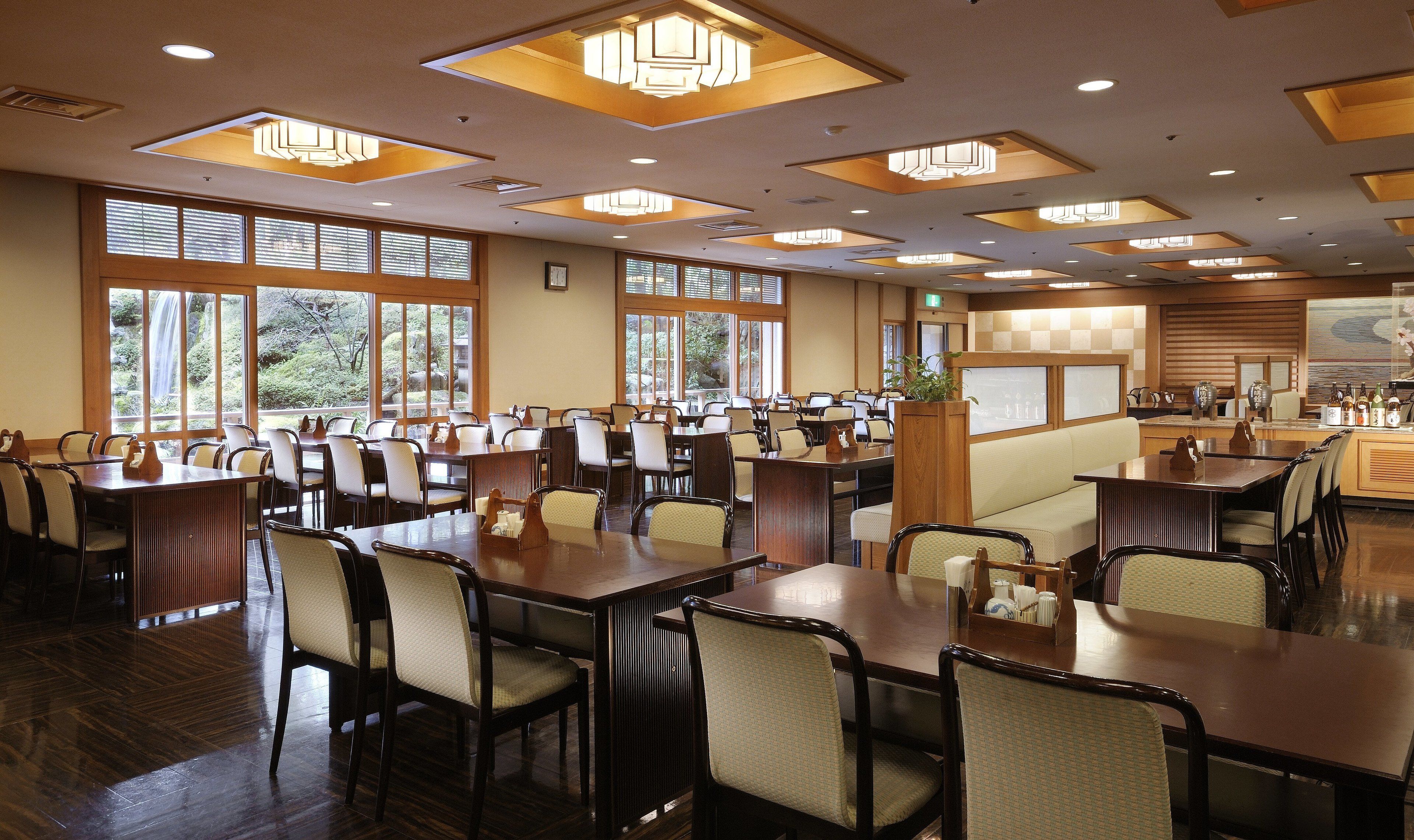 Un vaste espace dédié à la salle à manger, avec de nombreuses tables et chaises, de grandes fenêtres offrant une vue sur la verdure, et une esthétique japonaise traditionnelle caractérisée par des éléments en bois et des paravents coulissants.