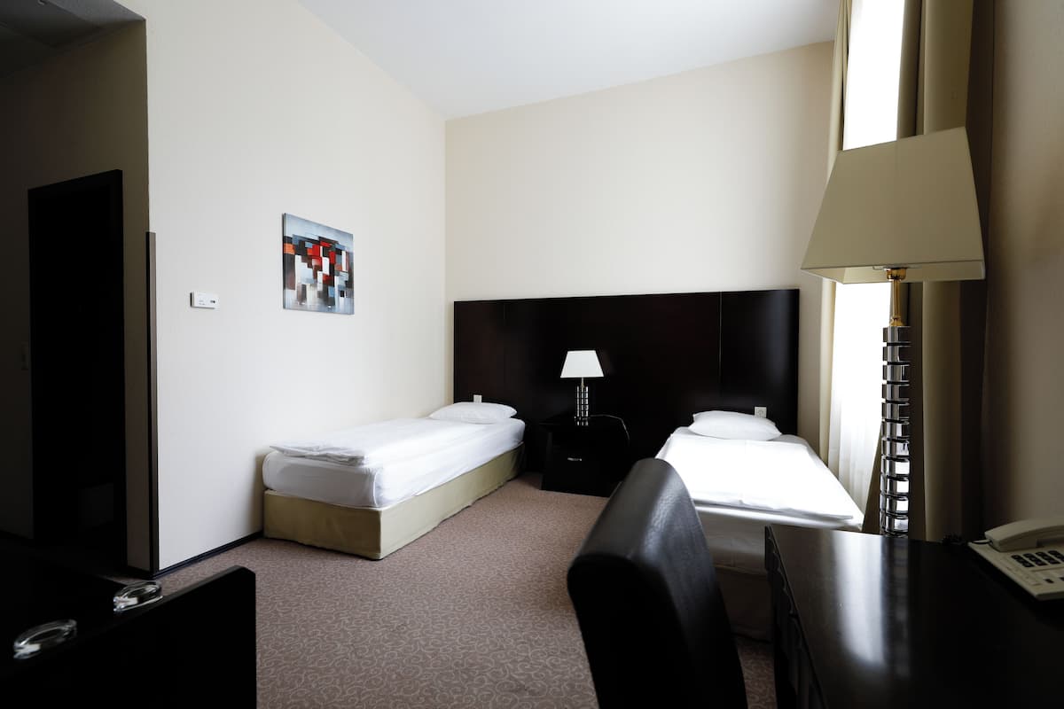Quarto de hotel com duas camas de solteiro, uma escrivaninha, uma cadeira, um abajur, uma TV e um quadro na parede.