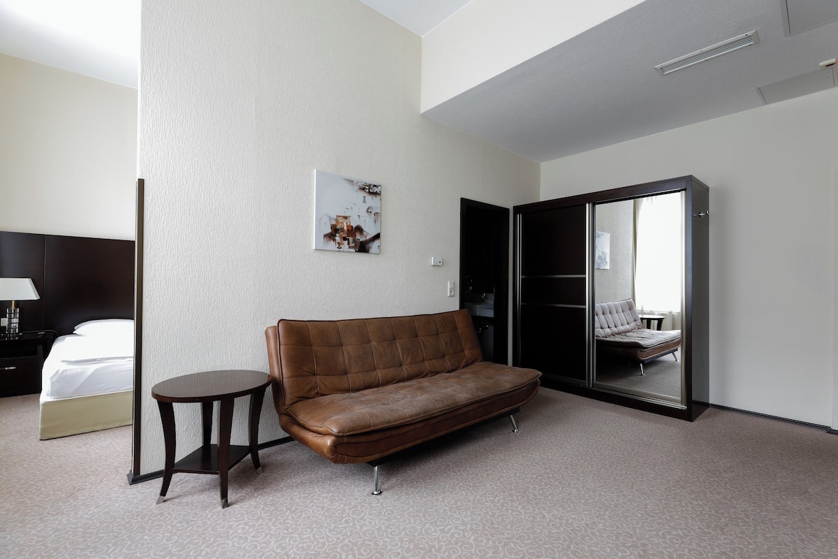 Quarto de hotel com sofá de couro marrom, uma mesinha redonda, cama com roupa de cama branca, um espelho grande e uma imagem pendurada na parede.