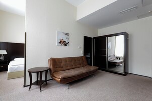 Habitación de hotel con sofá de cuero marrón, una mesita pequeña y redonda, cama con ropa blanca, un espejo grande y un cuadro colgado en la pared.
