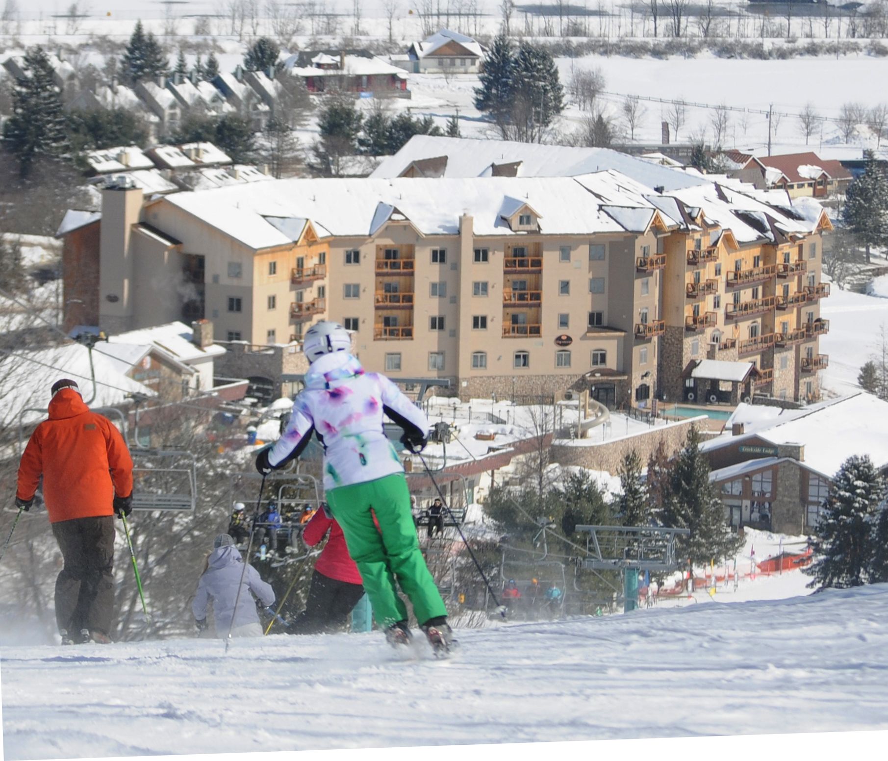 Une station de ski avec un grand bâtiment qui ressemble à un hôtel ou à un chalet, entouré d’arbres et de maisons couverts de neige.