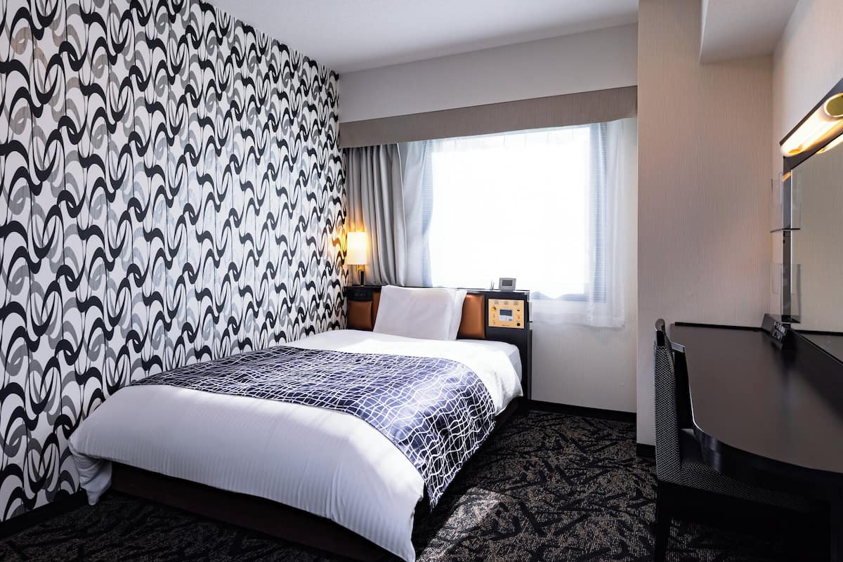 Habitación de hotel con papel tapiz estampado, cama con cubrecama estampado, mesita de noche con lámpara, escritorio con silla y una ventana grande con cortinas.